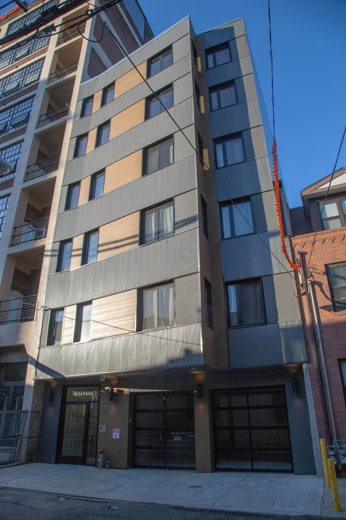 The exterior of the building is captured in strong natural light, showcasing a modern design with a combination of dark and light materials. Large windows punctuate the facade, providing a glimpse of the interior. A clean sidewalk leads toward the glass entrance.