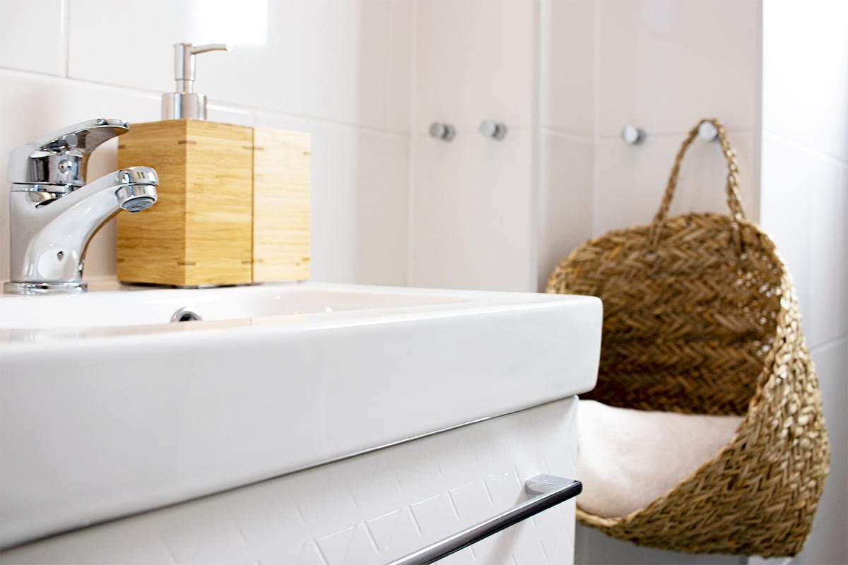 The bathroom features a modern sink with a sleek faucet and a wooden soap dispenser. A wicker basket is positioned nearby, providing decorative storage. Light reflects off the white tiles, enhancing the clean and functional layout.