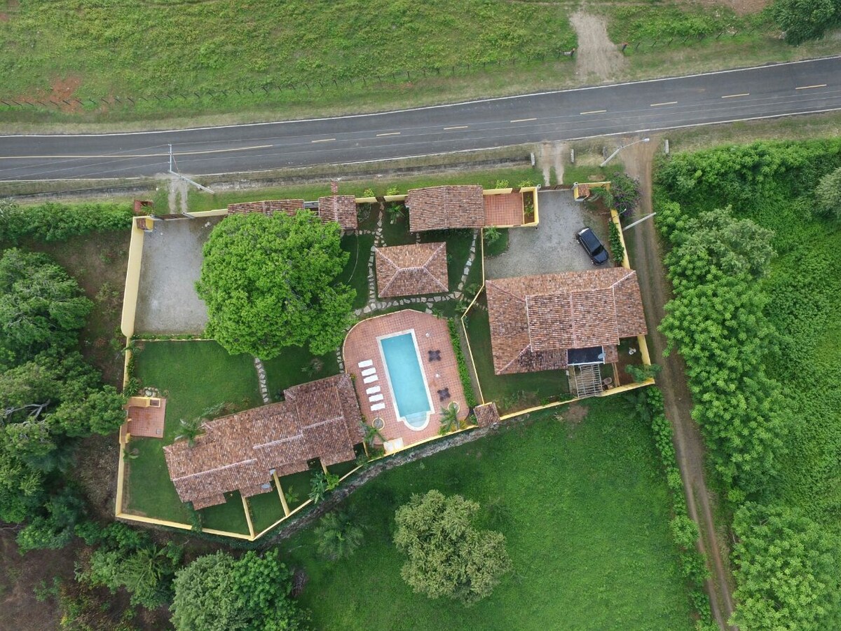 An aerial view captures the layout of the villa, highlighting the central swimming pool surrounded by lounge chairs. Lush greenery envelops the property, with multiple buildings featuring terracotta roofs positioned around the well-maintained garden area.