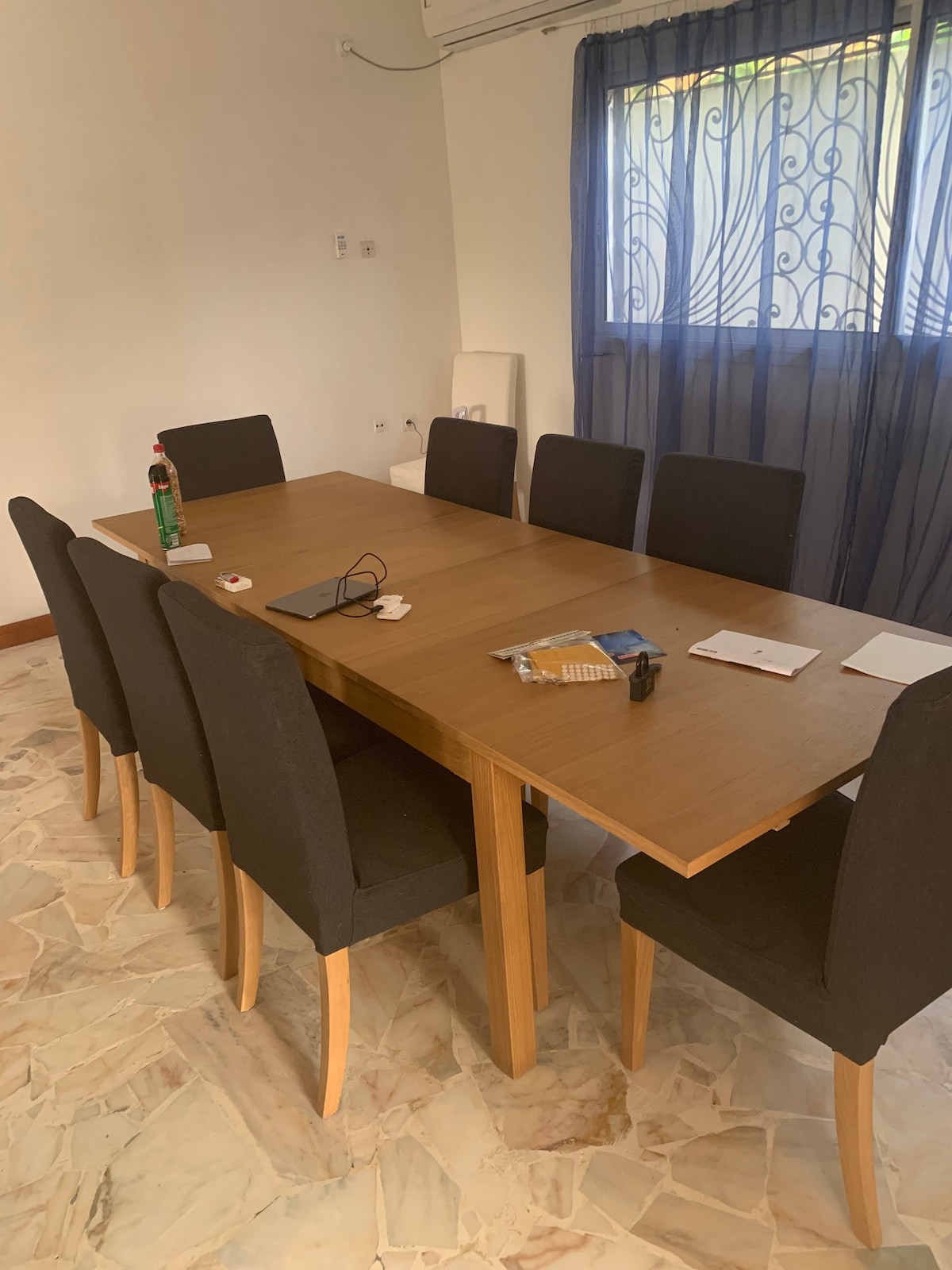 A wooden dining table is surrounded by six upholstered black chairs. An air conditioning unit is mounted on the wall, and natural light filters through a partially open window covered with sheer curtains. Small items are scattered across the table's surface.