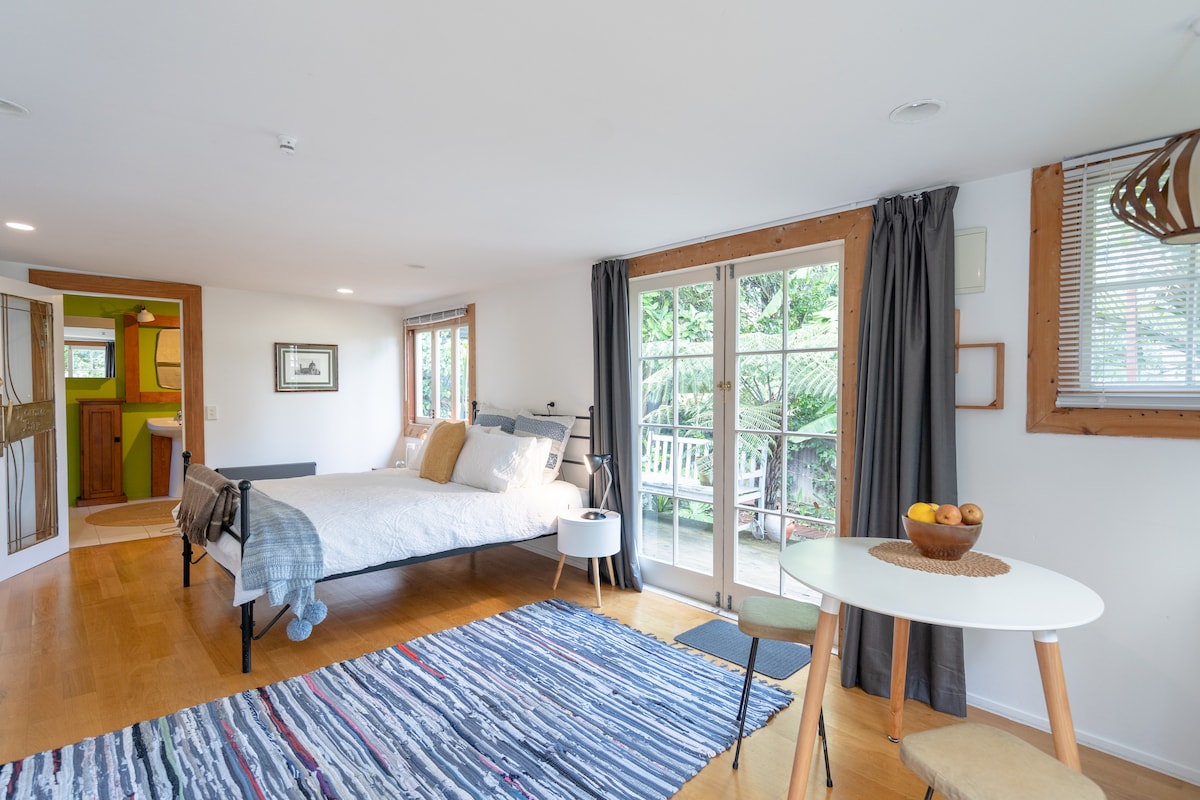 A bright studio space is equipped with a spacious bed and a round dining table. Sliding glass doors open to a garden view. Soft natural light highlights the wooden flooring and neutral wall tones. A soft area rug and contemporary furnishings create a cozy atmosphere.