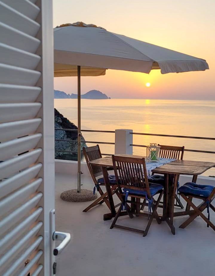 A terrace is shown, featuring a wooden table surrounded by folding chairs, under a large umbrella. The calm sea reflects the soft colors of the sunset, with distant islands visible on the horizon. The scene conveys a peaceful outdoor dining area.