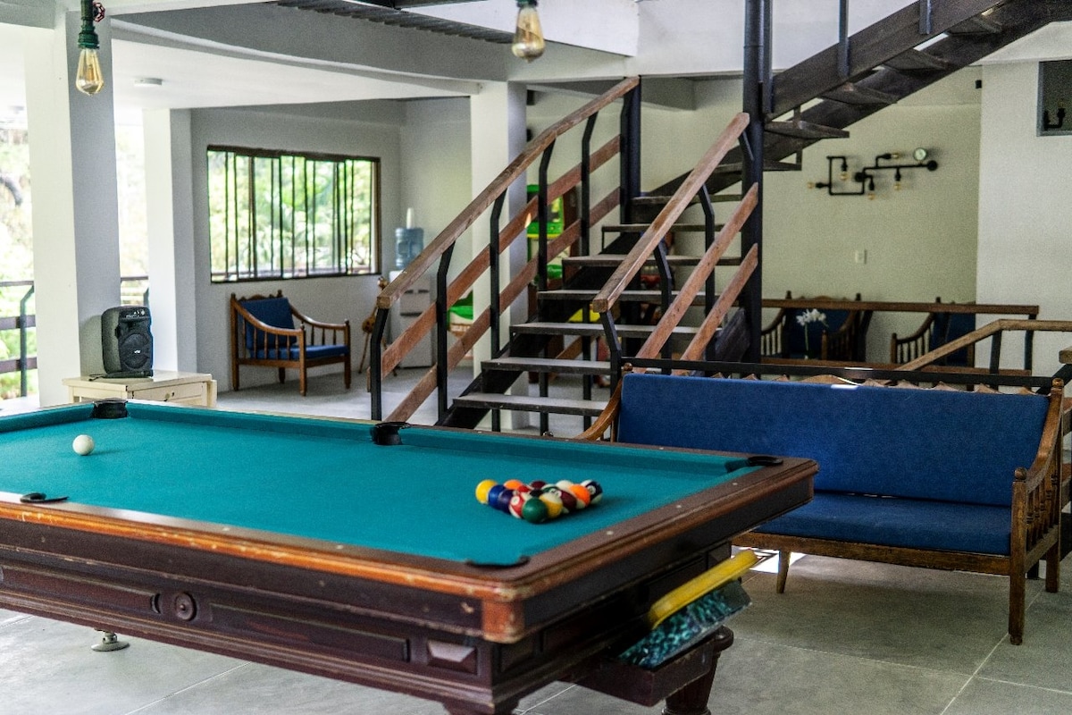 A shared recreational area is displayed, featuring a billiard table with a set of balls arranged for play. Comfortable seating options are visible, along with a staircase leading to an upper level. Natural light enters through large windows, contributing to a bright atmosphere.