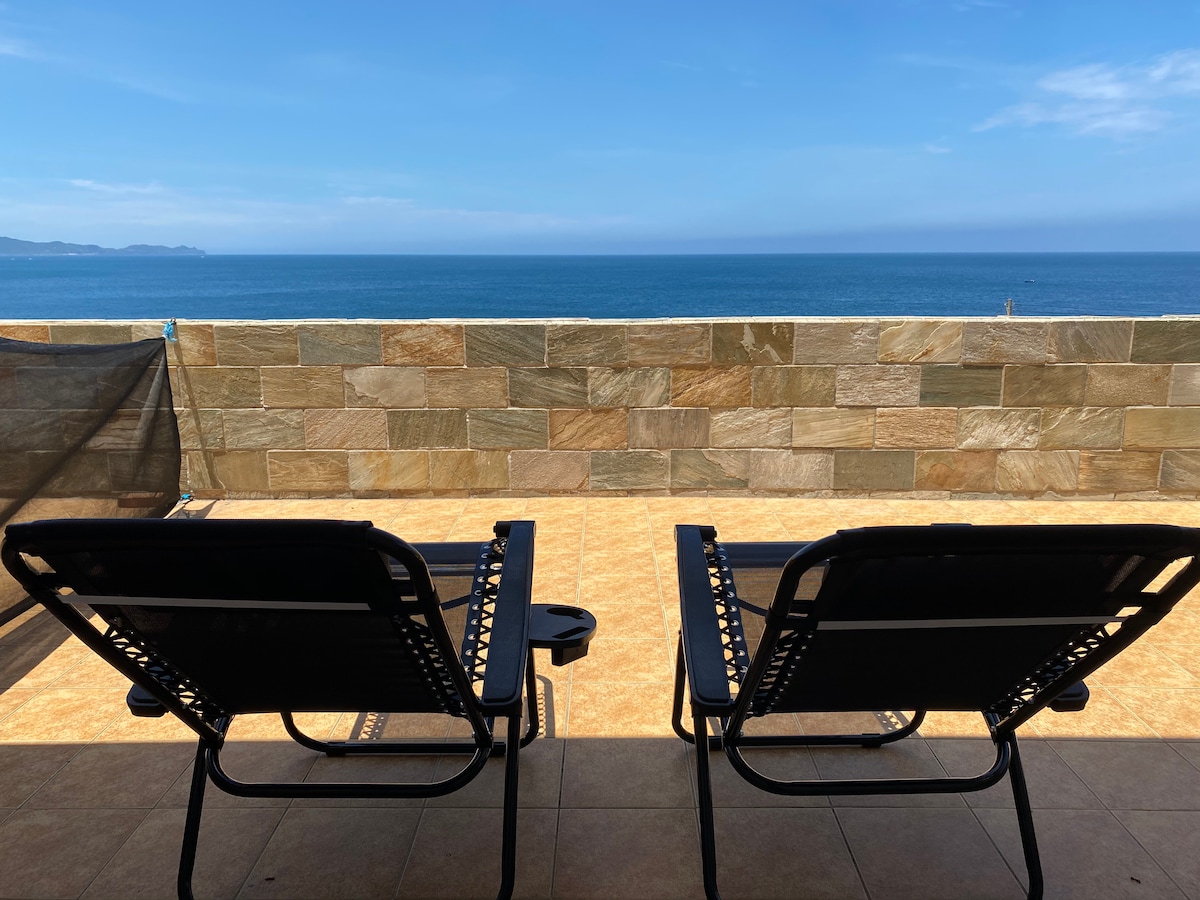 Two lounge chairs are positioned on a spacious patio, overlooking an unobstructed view of the ocean. The terrace features a natural stone wall, complementing the serene coastal landscape under a clear blue sky.