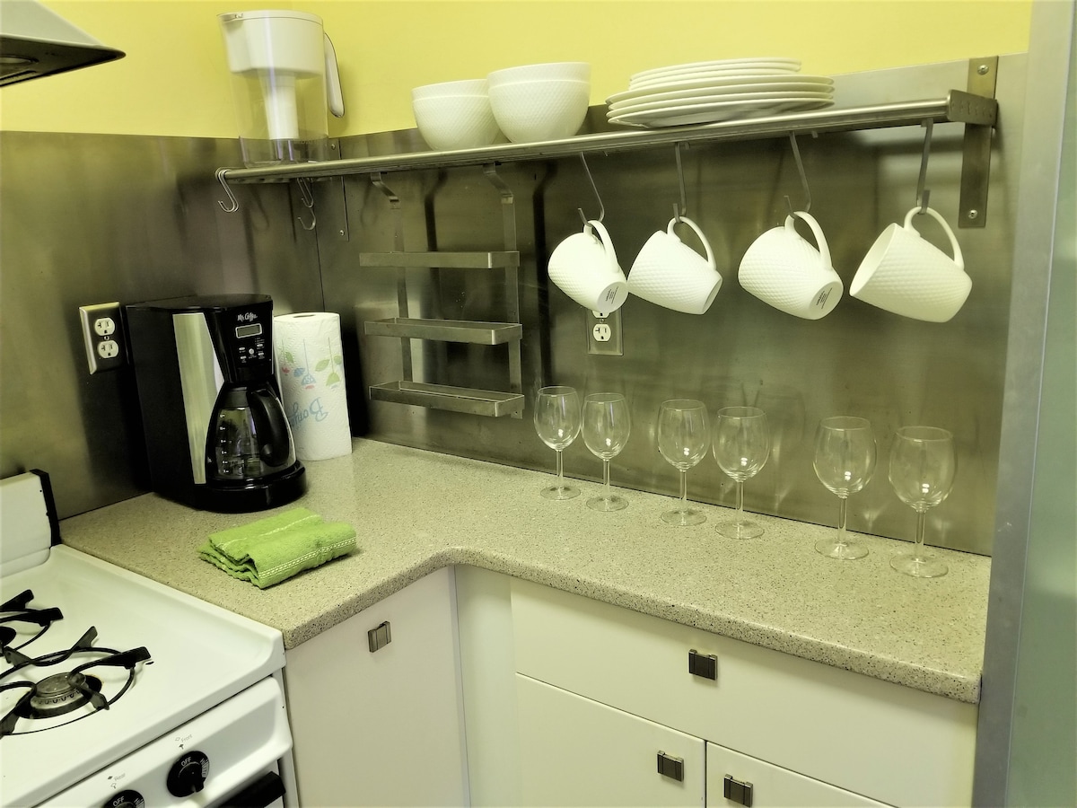 A compact kitchen area features stainless steel countertops and organized storage. Hanging cups dangle from a rack alongside a coffee maker. A set of six wine glasses rests neatly on the counter, complemented by stacked plates and bowls. A green dish towel is placed beside the sink.