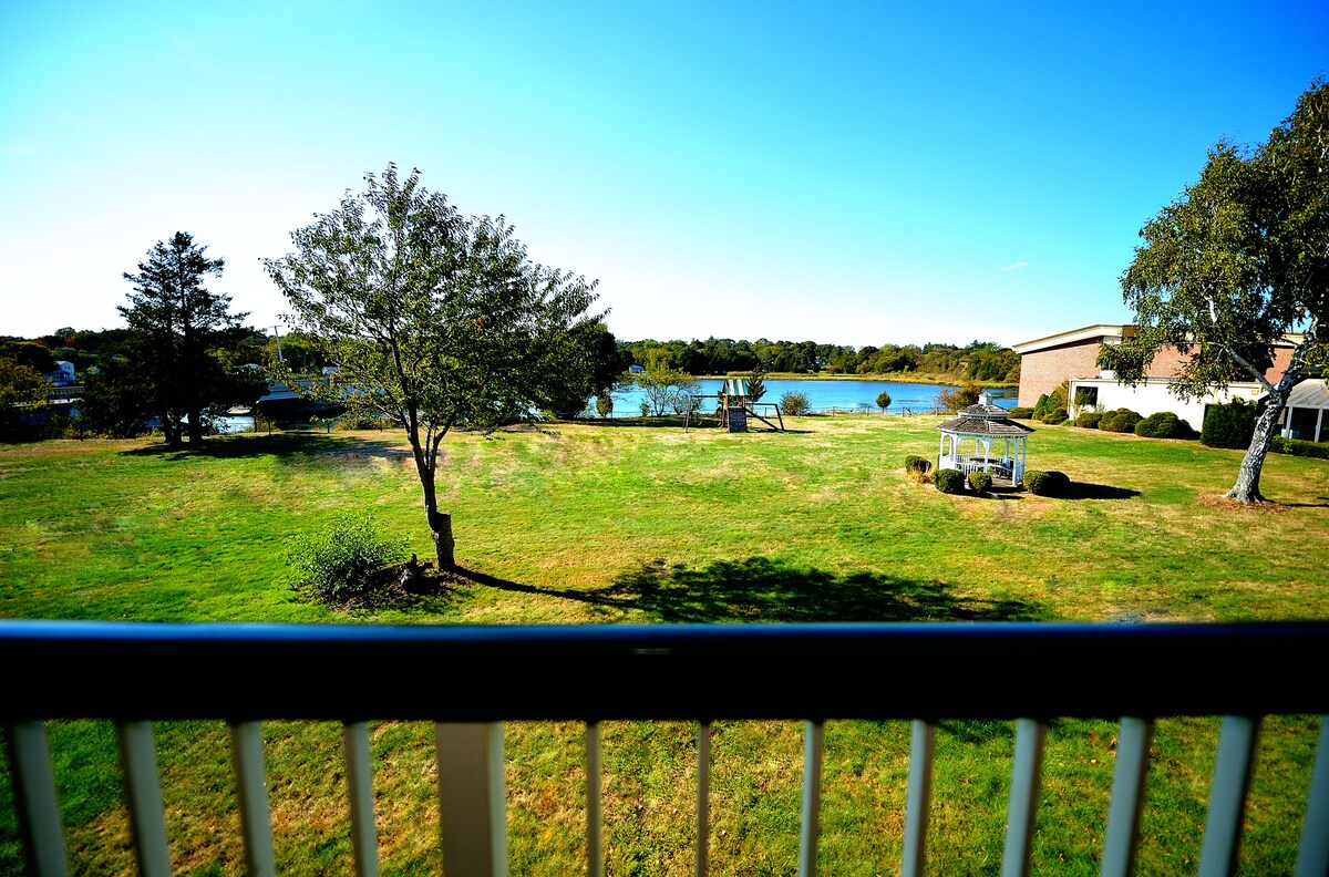 The image captures a serene waterfront view from a balcony, featuring a grassy area framed by trees. The Lee River is visible in the background, and a small seating area can be seen on the lawn, offering a tranquil outdoor space.