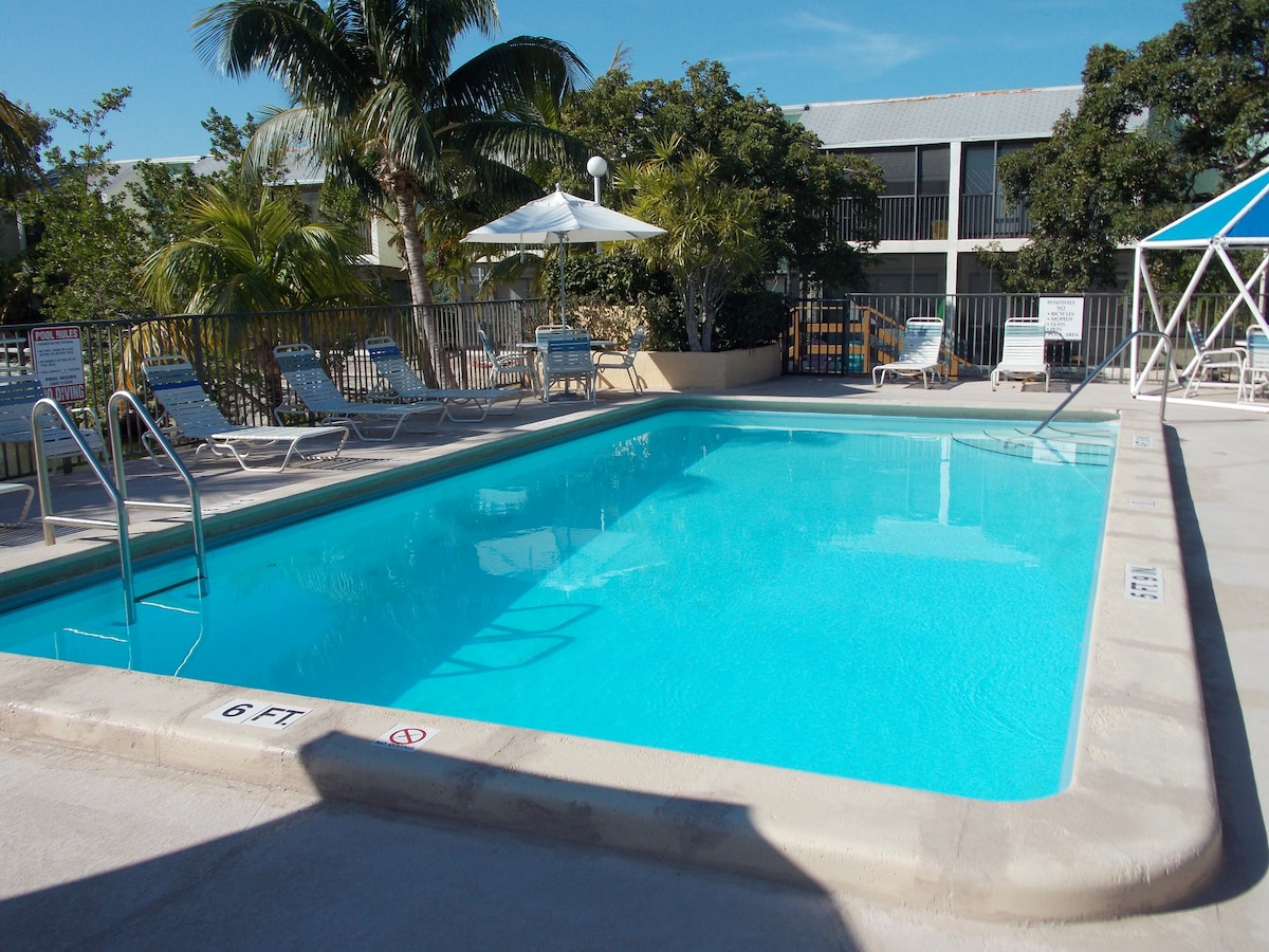 A refreshing swimming pool is surrounded by lounge chairs under palm trees. The clear blue water reflects the sunlight, inviting relaxation. A shaded area with a canopy is situated nearby, providing a comfortable retreat from the sun.
