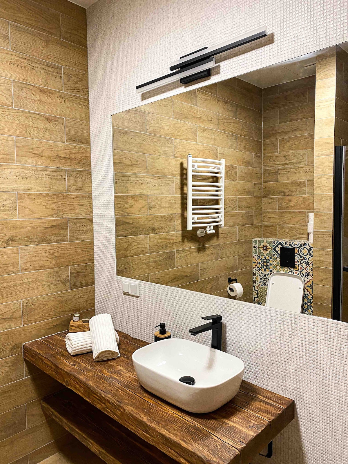 The modern bathroom features warm wooden tones and textured tiles. A large mirror reflects the area, which includes a stylish sink atop a wooden vanity. A heated towel rack and decorative elements add character, while essential toiletries are neatly arranged.