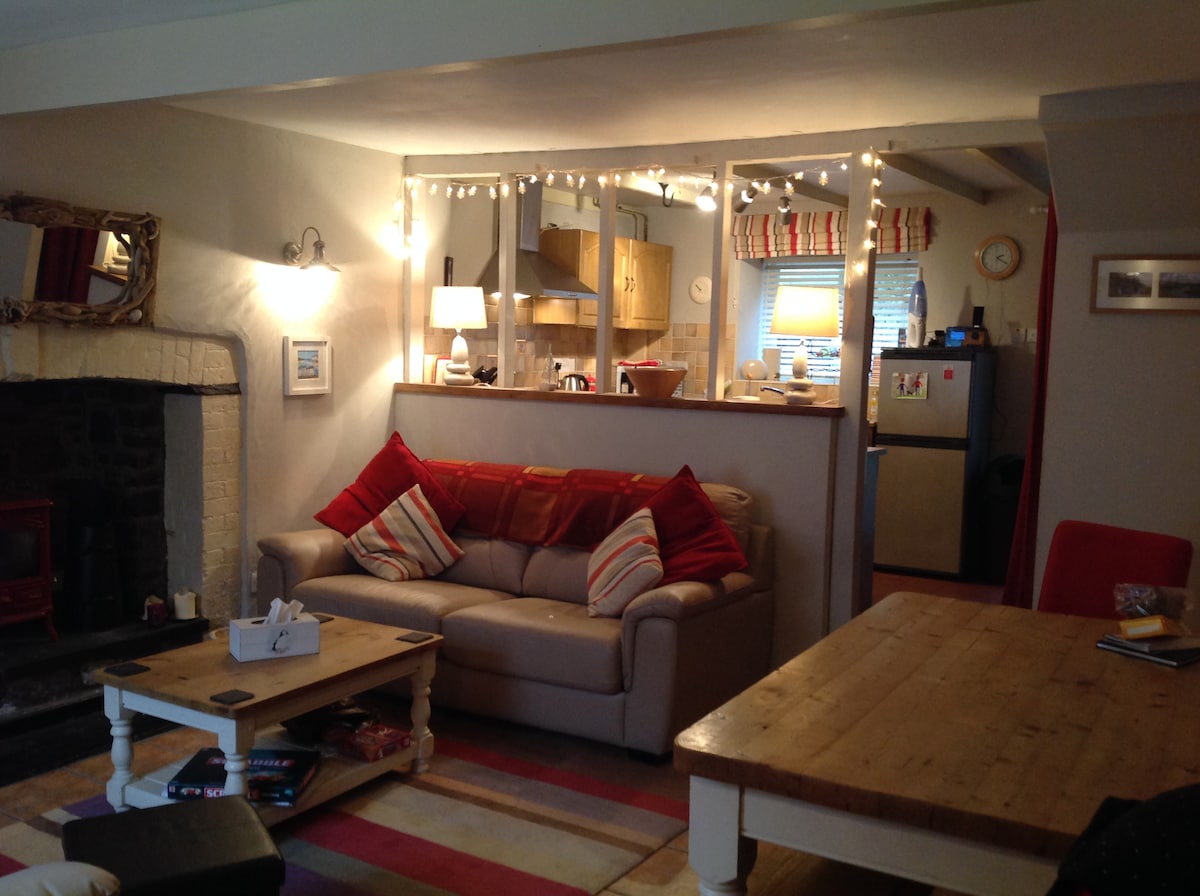 A cozy living area is visible, featuring a light beige couch adorned with red and white striped cushions. A wooden coffee table sits in front, with a patterned rug beneath. A stone fireplace provides warmth, and a small kitchen space is partially seen in the background.