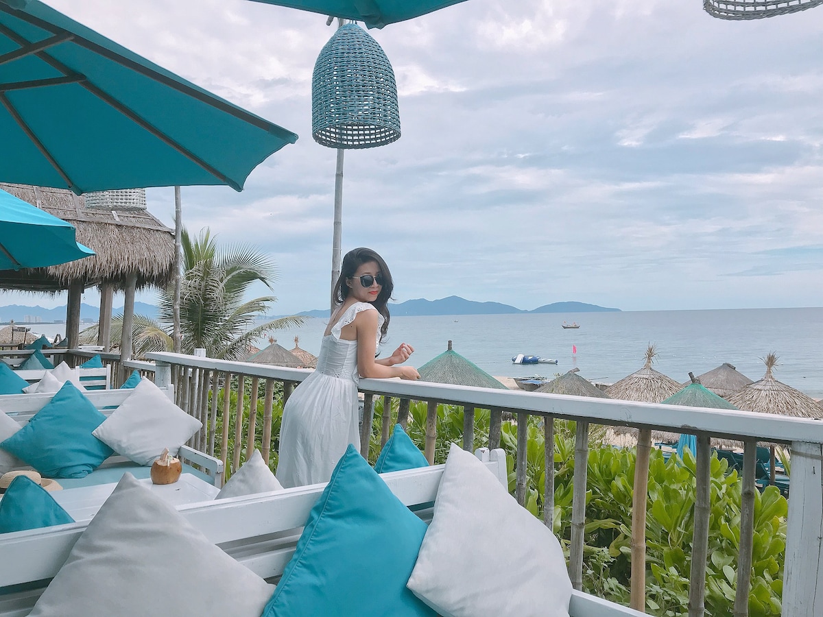A woman leans against a railing, overlooking a serene beach scene. Colorful umbrellas provide shade for the seating area, which is adorned with plush cushions. The ocean stretches out in the distance, framed by a few distant islands and a tranquil sky.