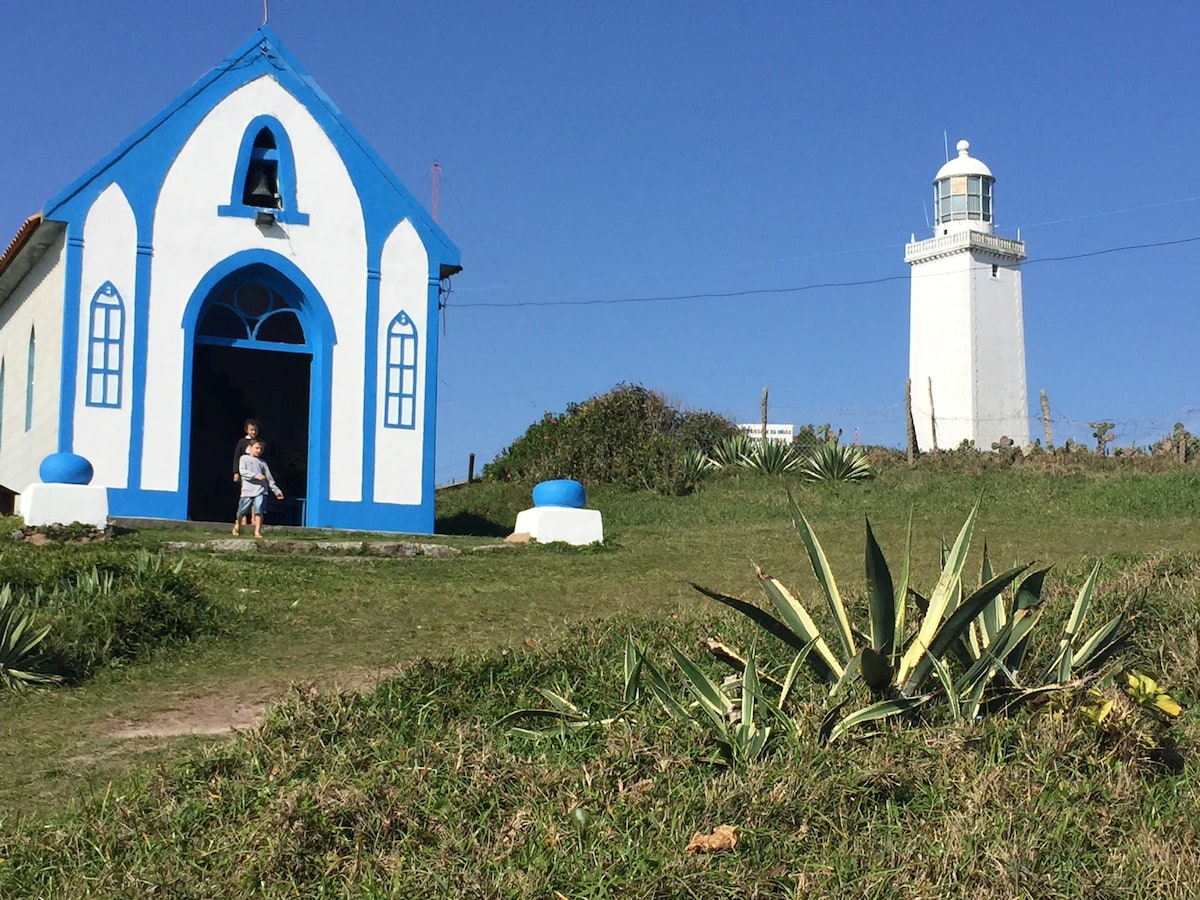 A white lighthouse stands prominently beside a small white and blue building, set against a clear blue sky. Green grass and agave plants are visible in the foreground, adding natural elements to the scene. A person is seen exiting the building.