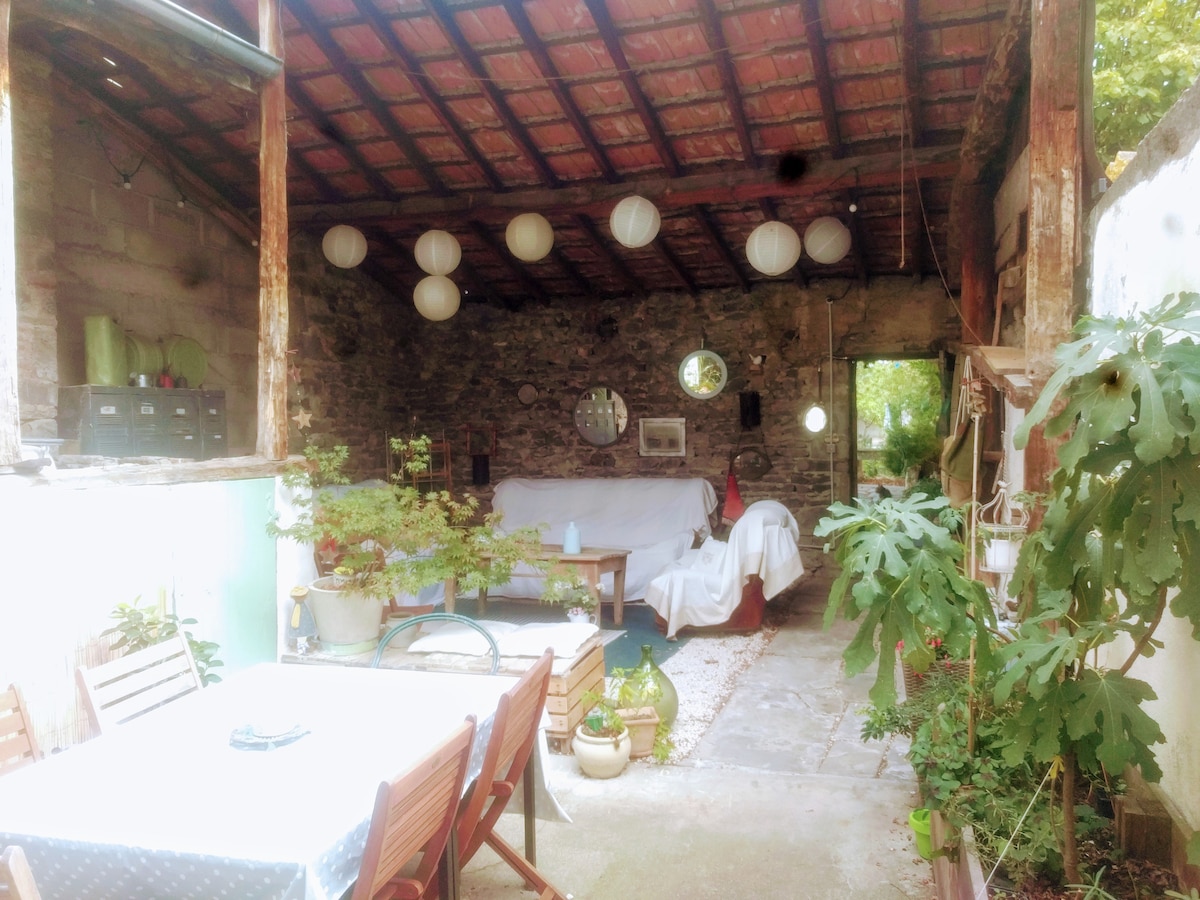 An open living space reveals a combination of natural stone walls and wooden beams, with soft lighting provided by decorative lanterns. Comfortable seating is arranged around a central area, surrounded by potted plants. A dining table with wooden chairs is positioned under a canopy of greenery.
