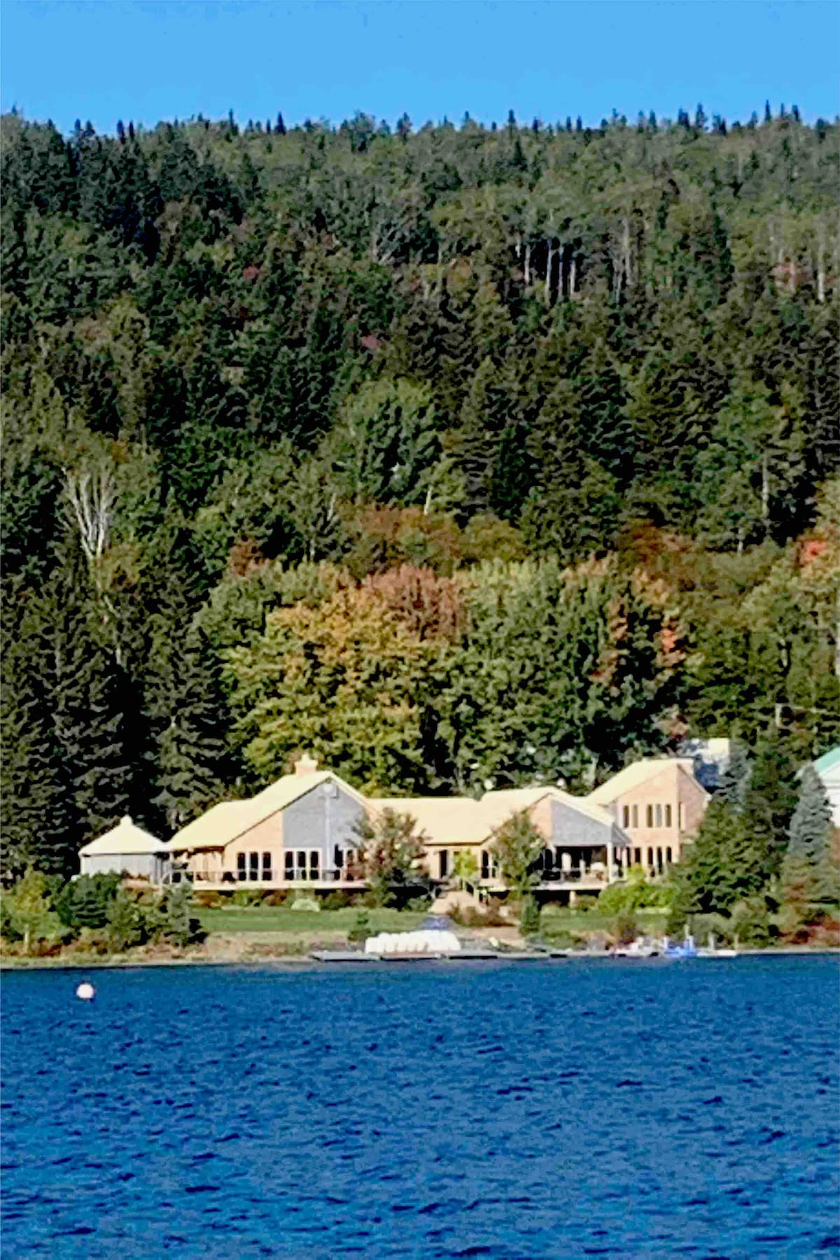 A spacious lakeside estate appears surrounded by vibrant greenery and dense forested mountains. The building's large windows reflect the natural surroundings, while a gentle shoreline is visible in the foreground, enhancing the serene lakeside setting.
