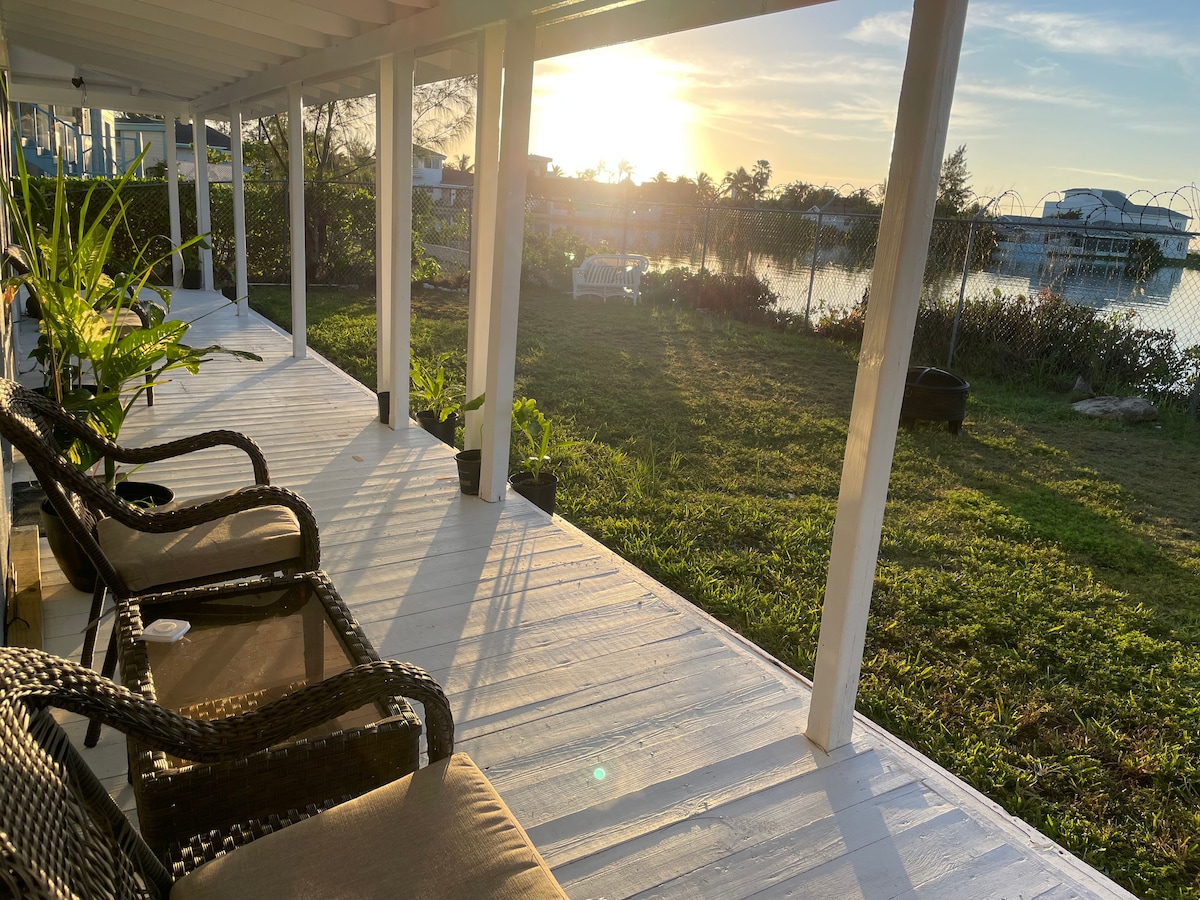 A wraparound patio features comfortable seating and is bordered by lush greenery. The sun sets over the tranquil water, creating a serene ambiance. Reflection of the golden light dances on the surface of the canal, enhancing the peaceful outdoor setting.
