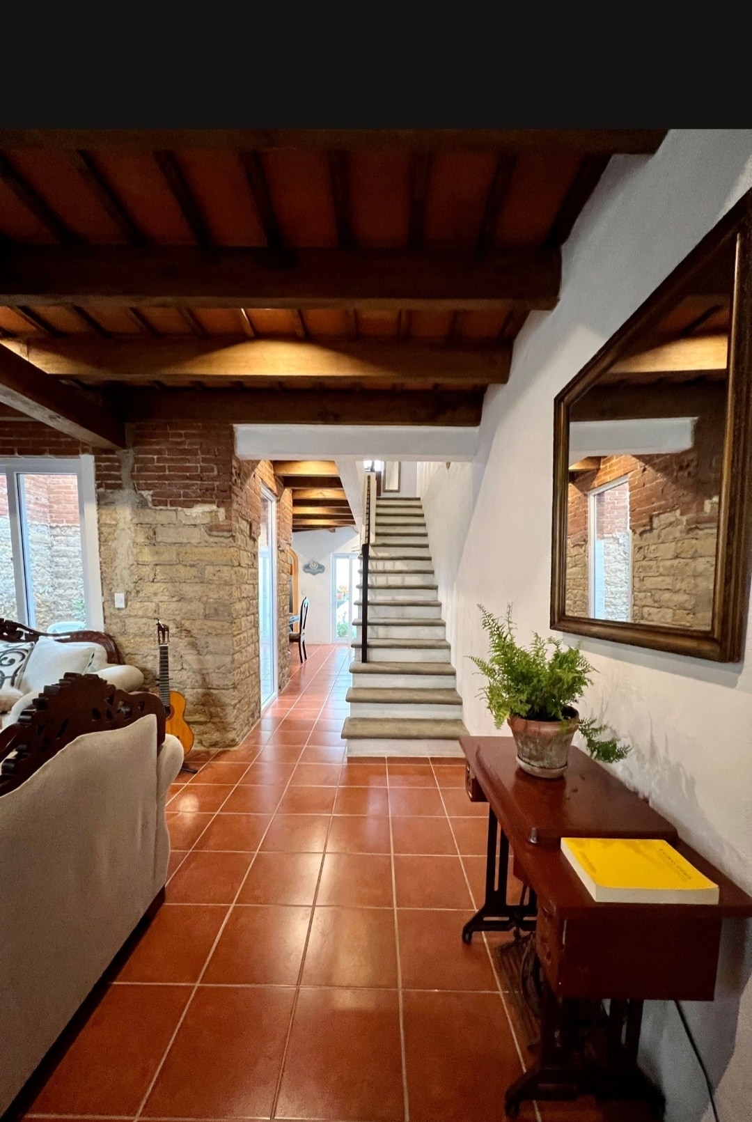 A hallway features a terracotta tiled floor and exposed wooden beams overhead. A wooden table with a potted plant is positioned against one wall, while a large mirror reflects the inviting space. Stairs lead to an upper level, illuminated by natural light from windows.