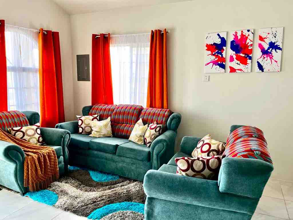 A living room features two plush sofas and a patterned armchair, arranged around a circular rug. Bright orange curtains frame the windows, allowing natural light to illuminate the space. Colorful artwork adorns the wall, adding a lively touch to the decor.
