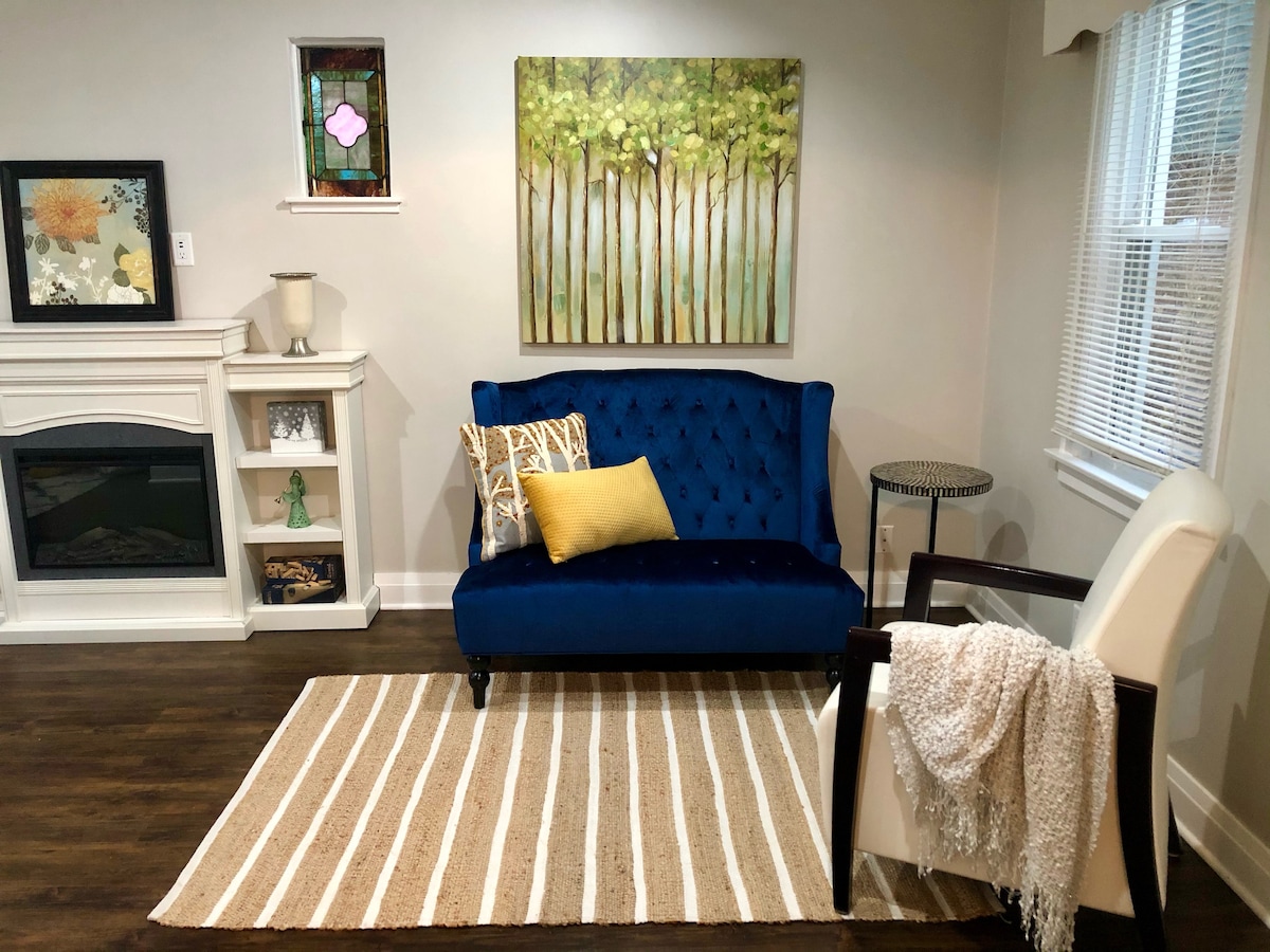 A cozy living area is highlighted by a plush navy sofa adorned with a yellow accent pillow. A light-colored area rug with stripes lies beneath. The room features a decorative painting of trees and a stained glass window, adding character to the inviting space.