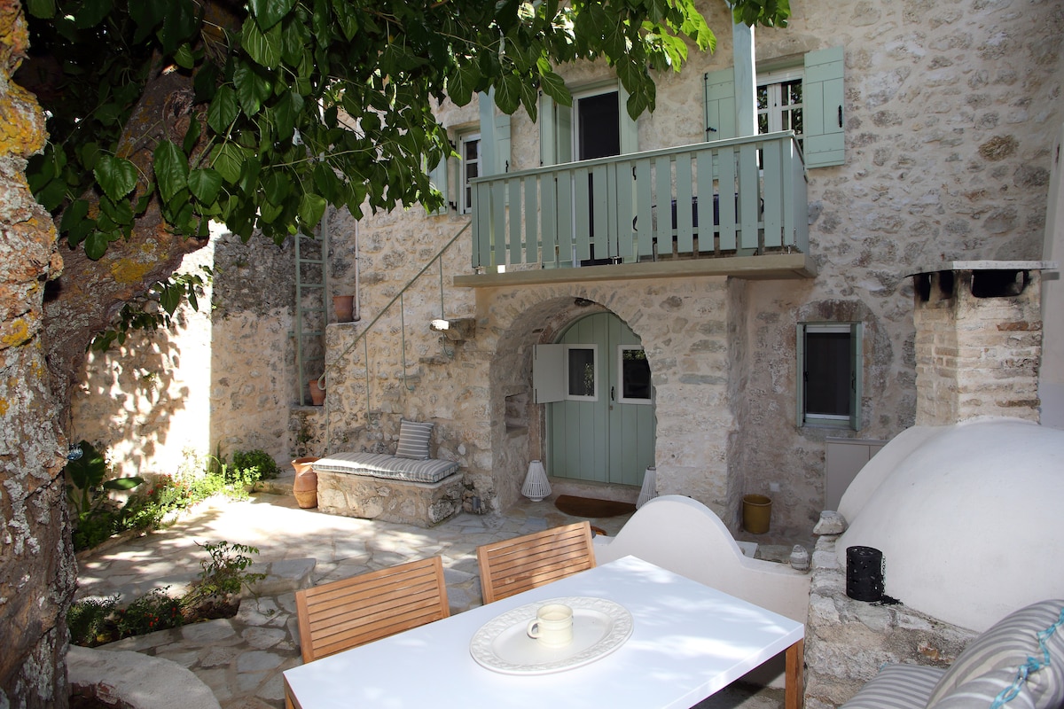 A charming outdoor space is revealed, featuring stone walls and a patio area with a white dining table. Comfortable seating is arranged nearby under lush greenery, providing shade. An arched entrance leads to the interior, with a set of stairs visible in the background.