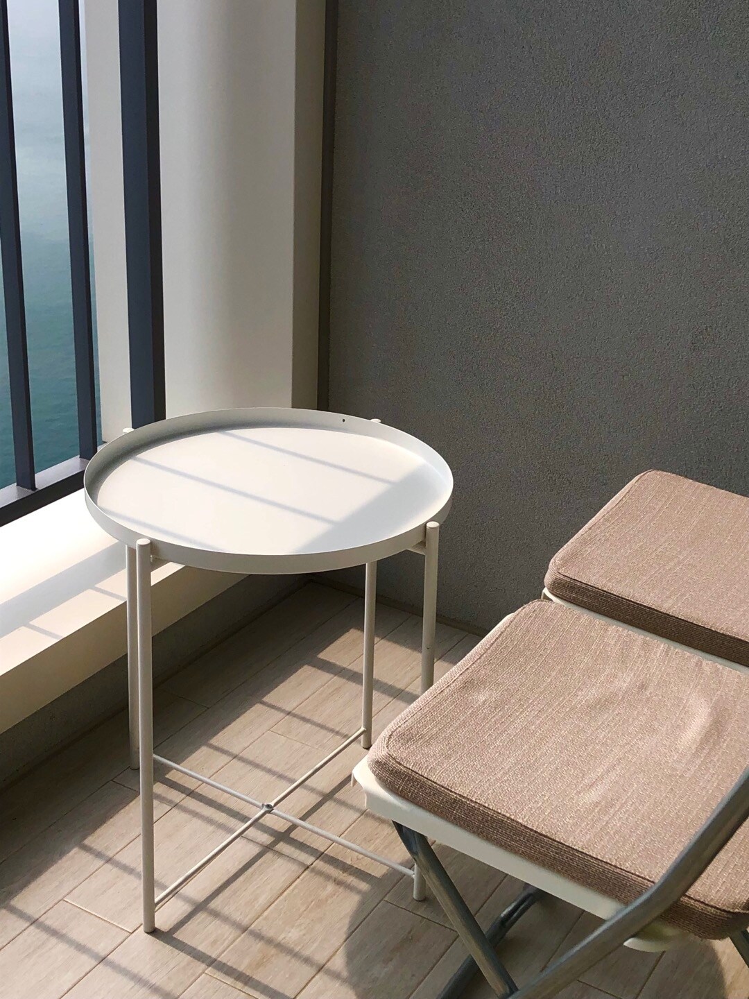A small round side table is positioned next to a cushioned chair on a balcony. Natural light filters through the large window, reflecting off the ocean visible beyond the railing. The serene atmosphere offers a space for relaxation.