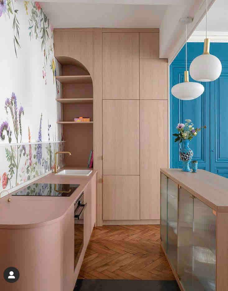 A modern kitchen features soft pink cabinetry with a minimalist design, complemented by floral wallpaper. Open shelving and a sink are seamlessly integrated. Two pendant lights hang above a central counter, while a decorative vase with flowers adds a touch of color to the space.