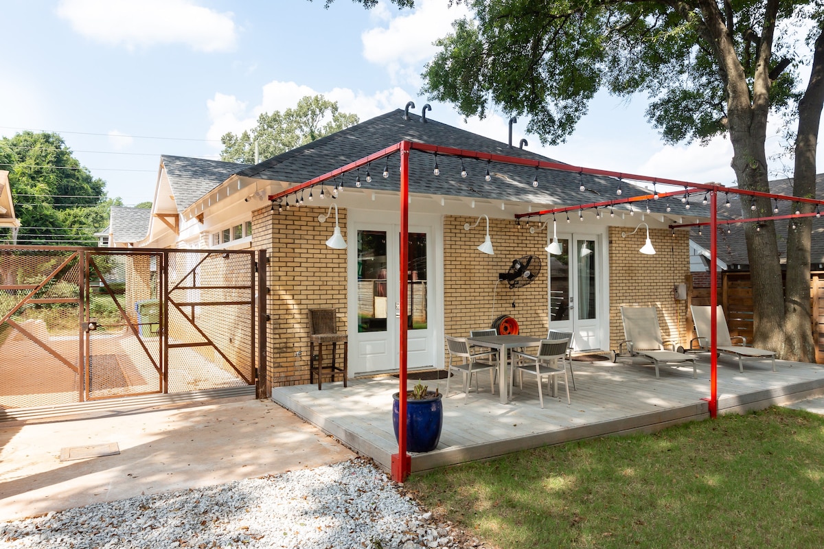 An outdoor seating area is positioned under a covered porch, featuring a dining table and chairs. Decorative lights hang overhead, illuminating the space. The area is bordered by a gated entrance, with a lush green lawn and trees in the background.