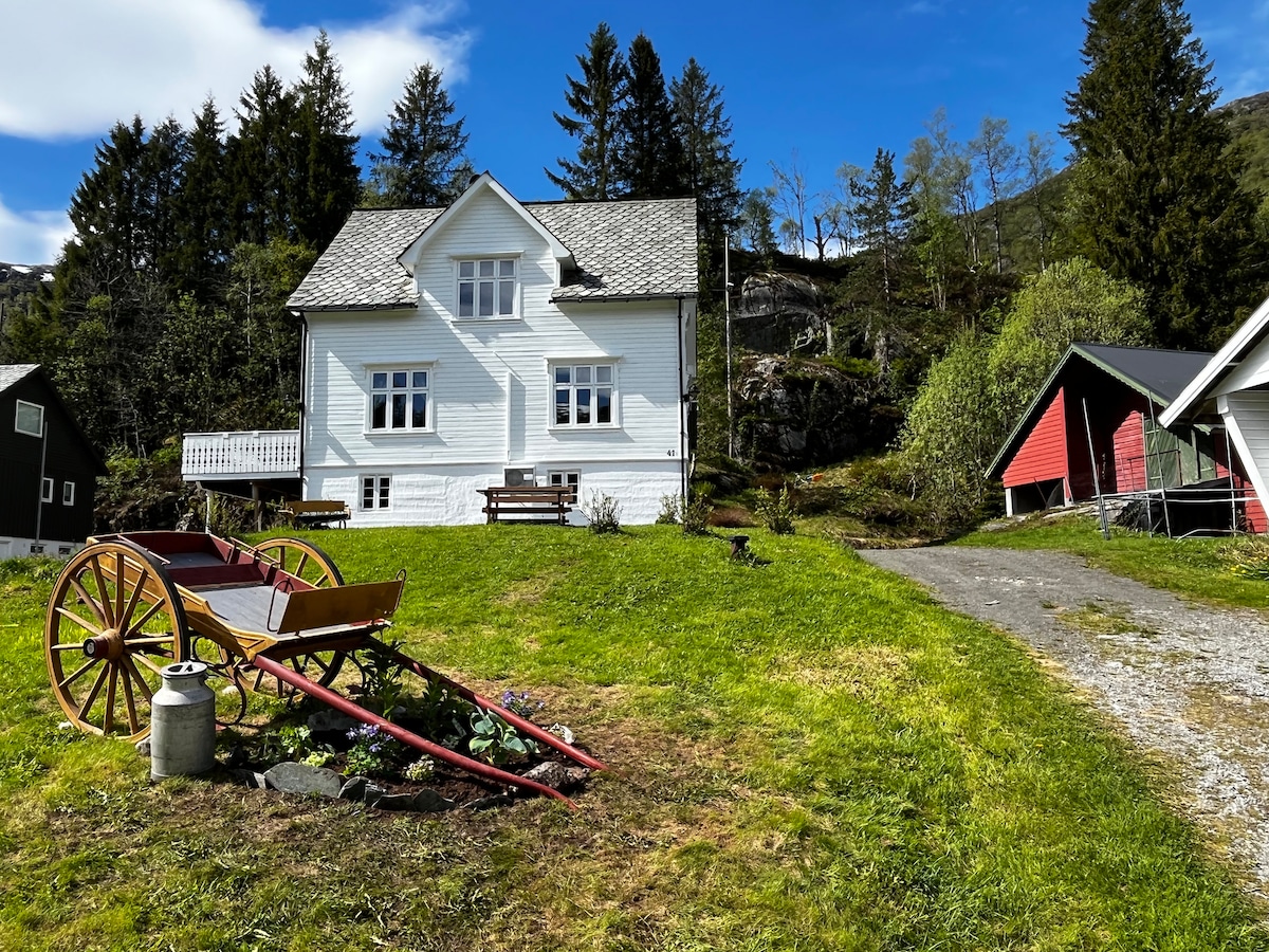 A traditional timbered farmhouse is displayed, surrounded by lush green grounds. The structure features large windows and a welcoming porch. A vintage wooden cart is positioned in the foreground, accompanied by a small stone water basin. Nearby are several other buildings, nestled among trees and a blue sky.