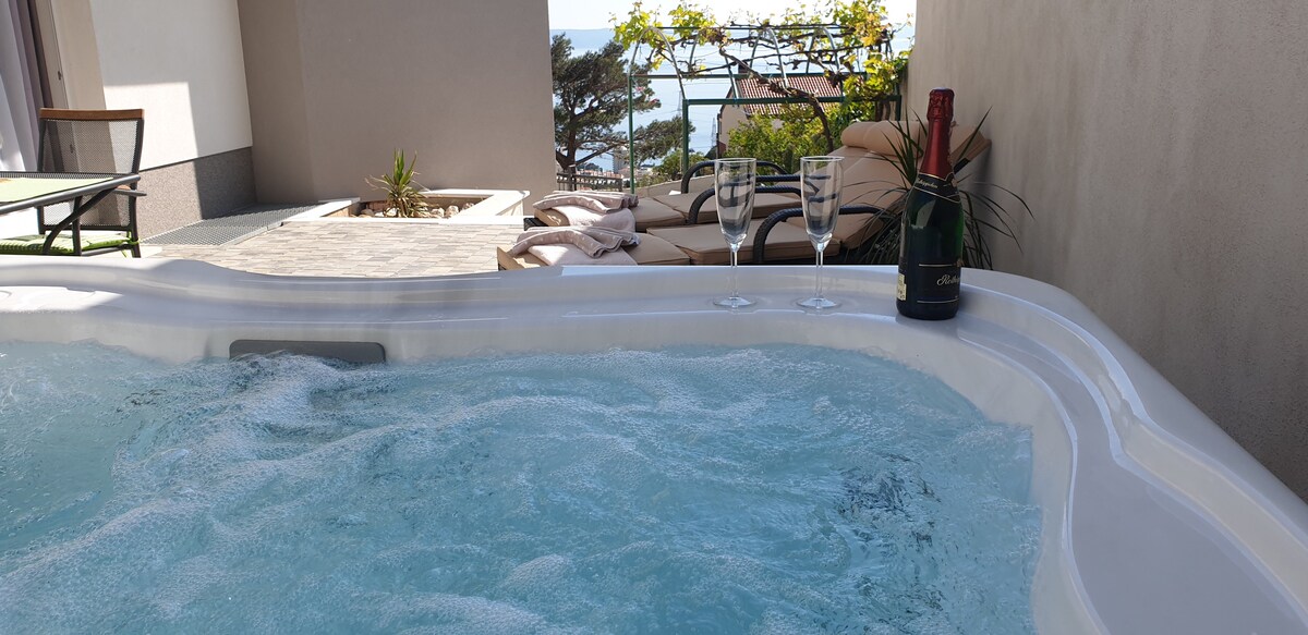 A hot tub is set on a terrace, surrounded by seating options and greenery. Glassware is placed nearby alongside a bottle. The area offers a view of the outdoor space, hinting at a tranquil atmosphere ideal for relaxation.