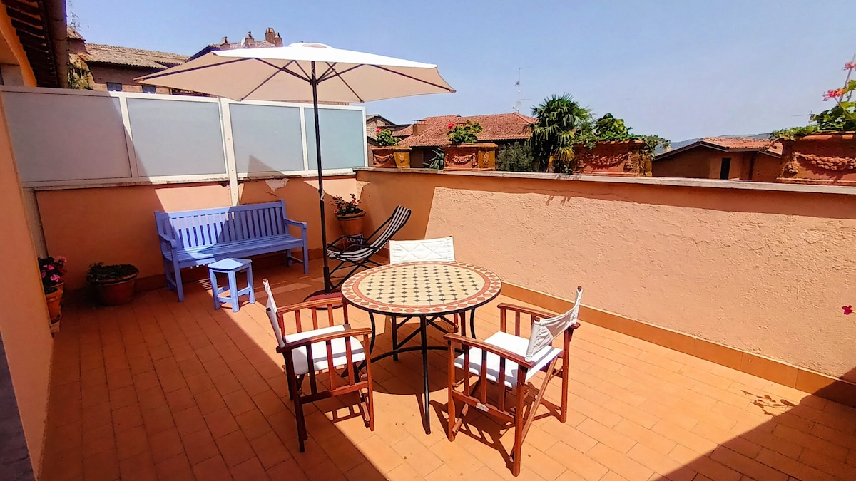 An expansive terrace is presented, featuring a round table surrounded by four wooden chairs. A blue bench and a lounge chair are included for relaxation. An umbrella provides shade, and colorful plants are arranged along the walls, enhancing the outdoor space.