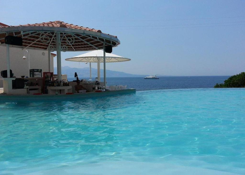 A serene infinity pool reflects the clear blue sky and the sparkling Ionian Sea. A bar under an elegant pergola offers seating and a striking view of the water. The distant coastline is visible, enhancing the tranquil setting of the pool area.