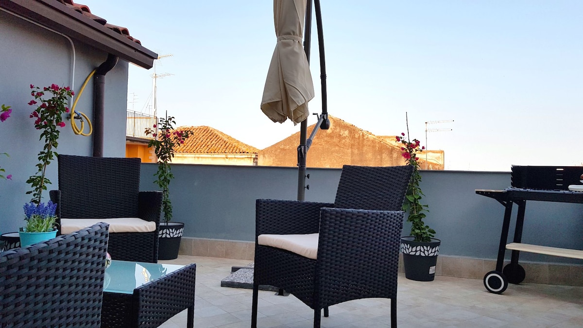 A spacious terrace is furnished with a seating area featuring four wicker chairs and a glass table. Potted plants add greenery to the space, while an umbrella provides shade. A barbecue grill is positioned nearby, enhancing the outdoor experience.