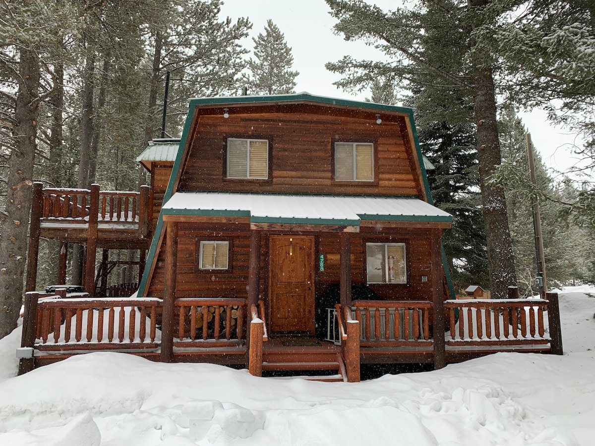 The cabin's exterior features a rustic wooden façade, with a green metal roof and multiple windows. Snow blankets the ground, while tall trees surround the property, creating a serene winter setting. A wrap-around deck is visible, enhancing the cabin's charm.