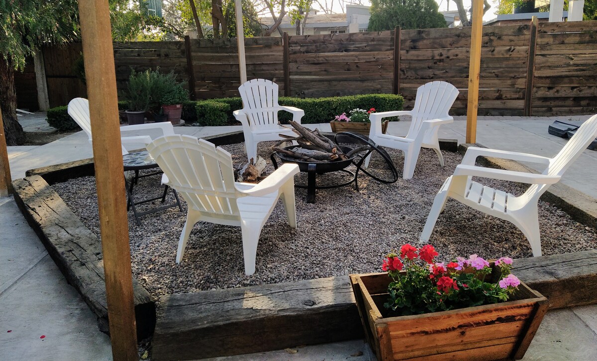A cozy seating area is arranged around a fire pit, surrounded by gravel and wooden borders. Five white Adirondack chairs provide comfortable seating, while colorful flowers bloom in a nearby planter. The space is designed for relaxation and social gatherings.