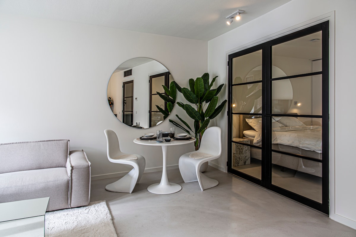 A light-filled living area is presented with a circular mirror and a stylish round dining table surrounded by white chairs. A large indoor plant adds a touch of greenery, while sliding glass doors lead to the bedroom, softly illuminated by warm lighting.