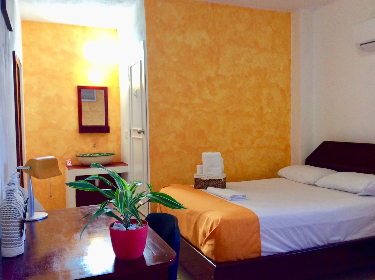 A bright room features warm yellow walls and a spacious bed with clean sheets. A desk with a lamp is positioned beside a small plant. The private bathroom is accessed through a door, and natural light is provided by a ceiling fixture.