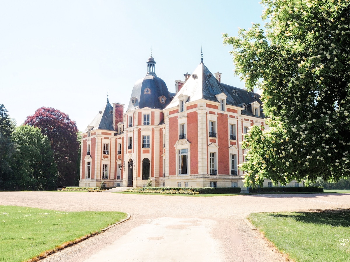 A grand château with distinctive architecture is portrayed, featuring elegant red brick walls and a prominent dome. Lush greenery surrounds the building, enhancing the serene setting. A well-maintained pathway leads to the entrance, inviting visitors to explore the grounds.