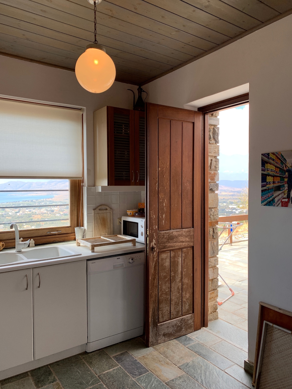 A kitchen space is visible, featuring a white sink and a dishwasher, with a microwave positioned nearby. A wooden door stands ajar, leading to an outdoor area with expansive views. Natural light enters through the window, illuminating the simple, functional design.