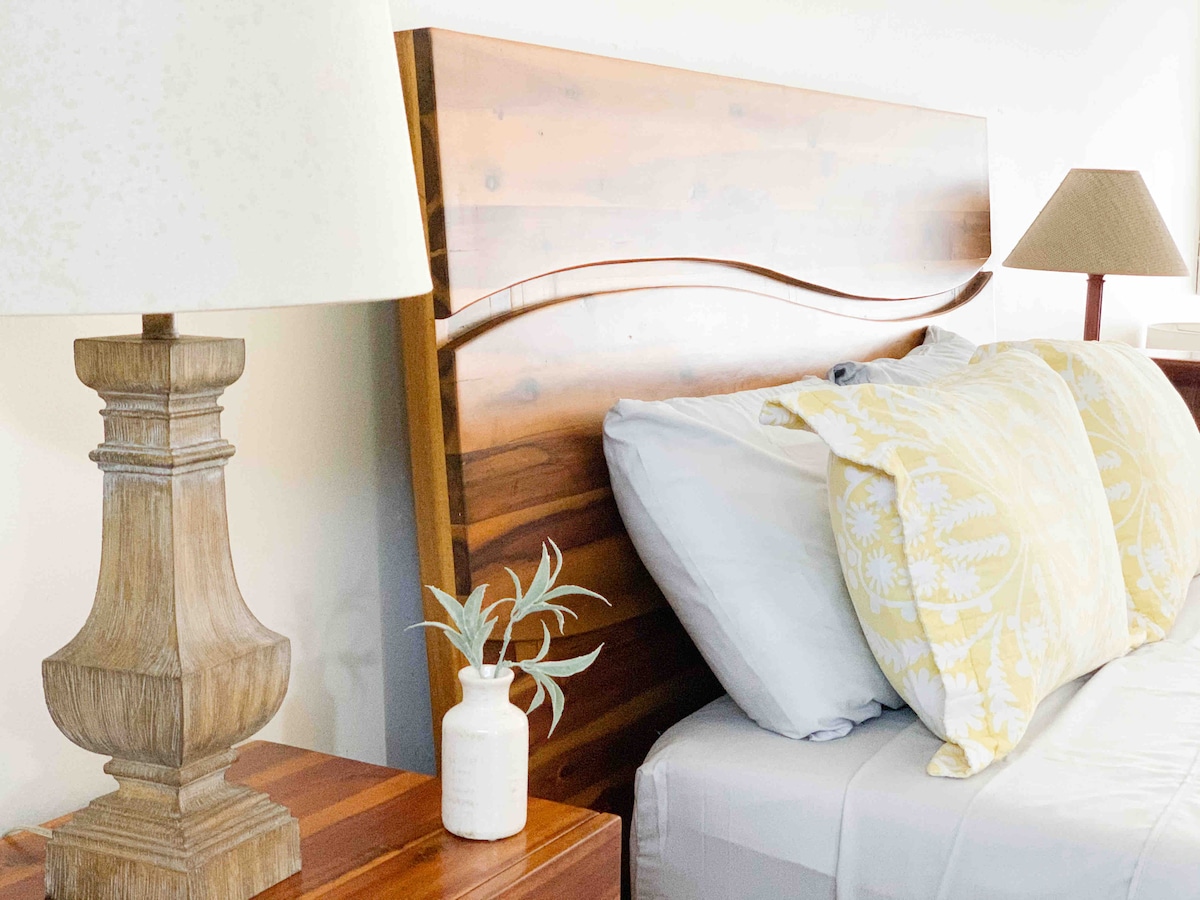 A cozy sleeping area is presented with a wooden headboard and two pillows adorned with a yellow floral pattern. A bedside table holds a decorative lamp and a small vase with greenery, contributing to a serene environment.