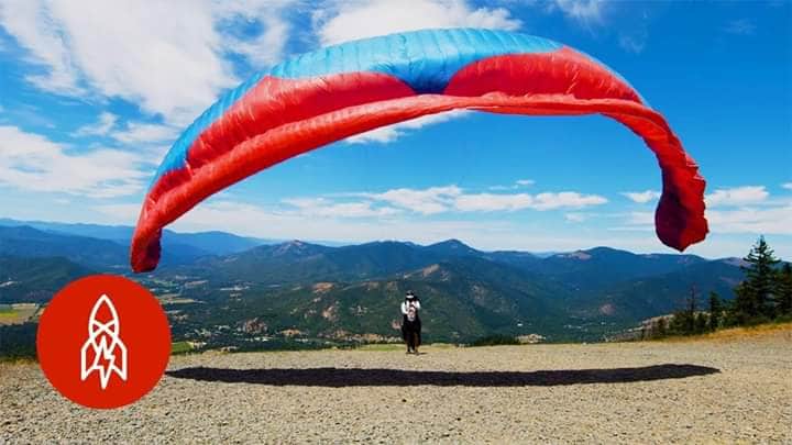A paraglider is prepared for flight, with a large red and blue wing spread wide above. The backdrop features expansive mountain views under a clear blue sky, while green trees line the area, creating a sense of adventure in nature.