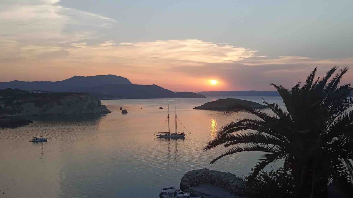 A serene view of Almyrida Bay is presented, featuring a calm sea reflecting the colors of sunset. Silhouetted palm fronds frame the image, while distant hills outline the horizon. A sailboat rests peacefully on the water, adding to the tranquil atmosphere.