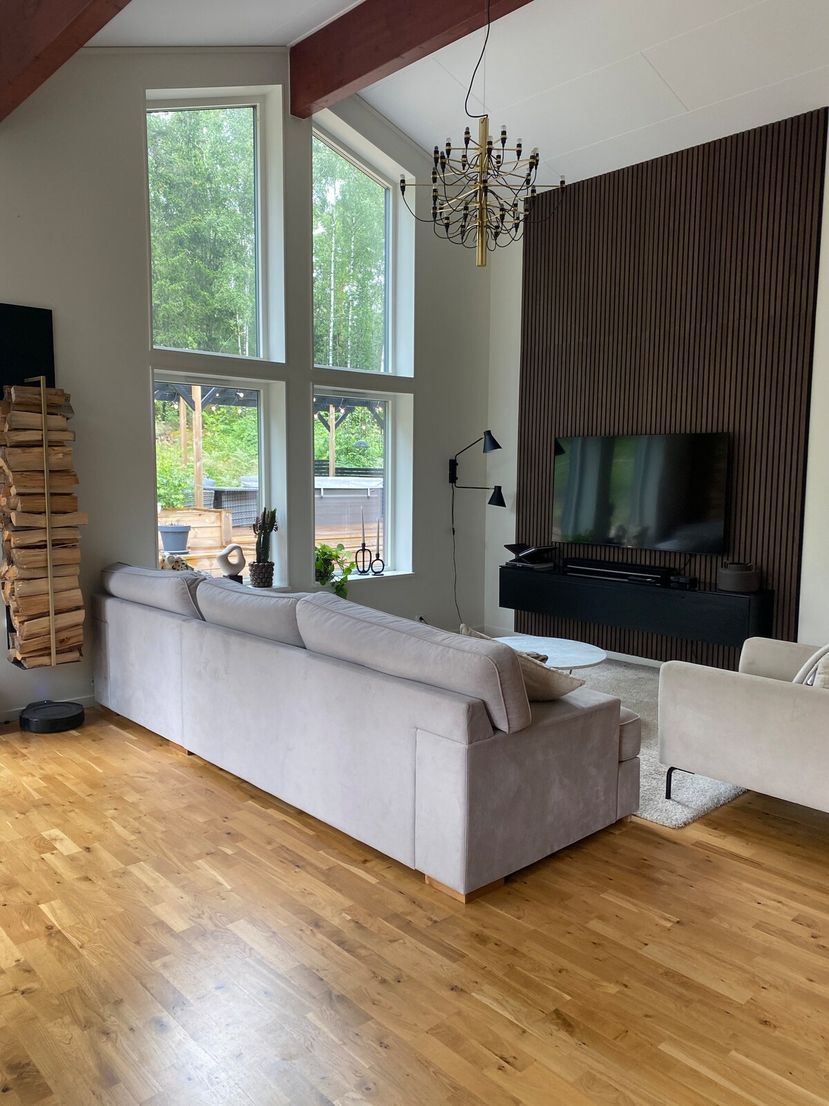 A spacious living area features tall windows allowing ample natural light. A modern gray sofa is positioned facing a wall-mounted television, complemented by a stylish black cabinet. A wooden accent wall adds texture, while a cozy chair and decorative plants create a welcoming ambiance.