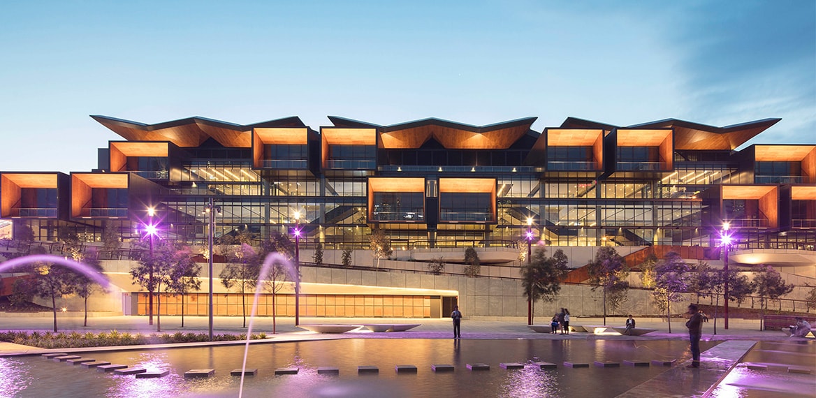 The modern building showcases a striking architectural design with angular roofs and large windows. Soft lighting illuminates the exterior, while water features create a serene atmosphere in the foreground. Trees and seating areas are present, enhancing the outdoor space.