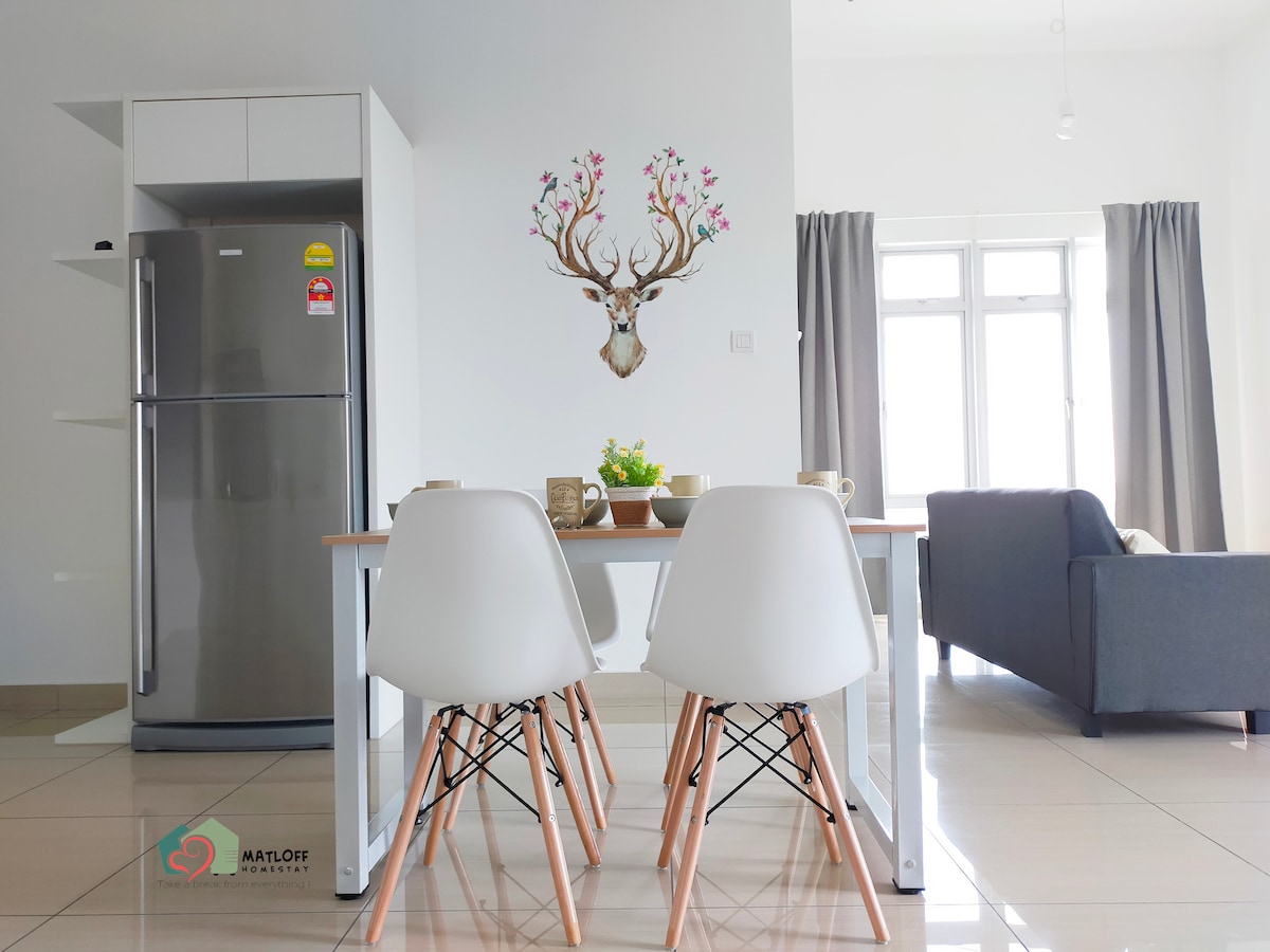 A modern kitchen area is highlighted by a sleek refrigerator and a dining table surrounded by two minimalist chairs. A decorative wall feature depicting a deer with antlers adds a unique focal point, while natural light fills the space through large windows, complemented by light gray curtains.