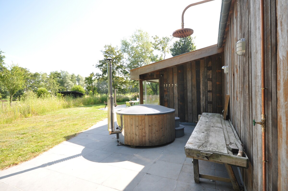 A wooden hot tub is situated on a stone patio, surrounded by a lush green landscape. A rustic outdoor shower is positioned nearby, with a wooden bench along the side, providing a serene space for relaxation in nature.