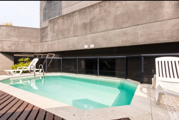 A swimming pool is set within an outdoor area, surrounded by a glass enclosure. Lounge chairs are positioned nearby on a wooden deck, and green plants are visible in the background, adding a touch of nature to the modern setting.