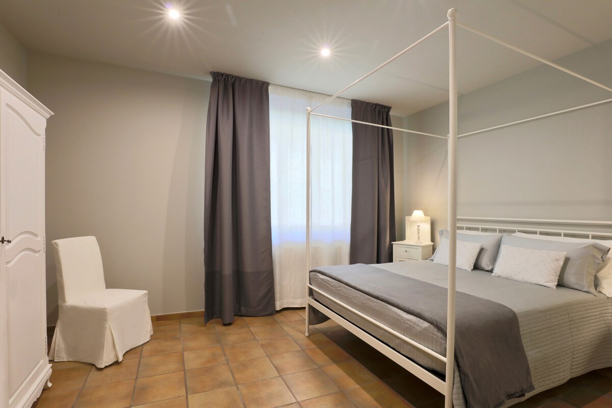 A spacious bedroom features a canopy bed with soft linens. Natural light filters through a large window, complemented by patterned curtains. A small white chair and a lamp beside a nightstand are present, enhancing the serene setting.