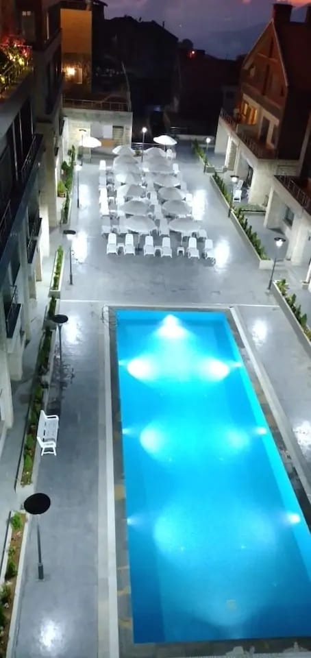 An aerial view captures a well-lit pool area featuring a clear blue swimming pool surrounded by comfortable lounge chairs and umbrellas. Soft illumination reflects off the water, enhancing the tranquil setting. Surrounding greenery and stone pathways create a welcoming atmosphere.