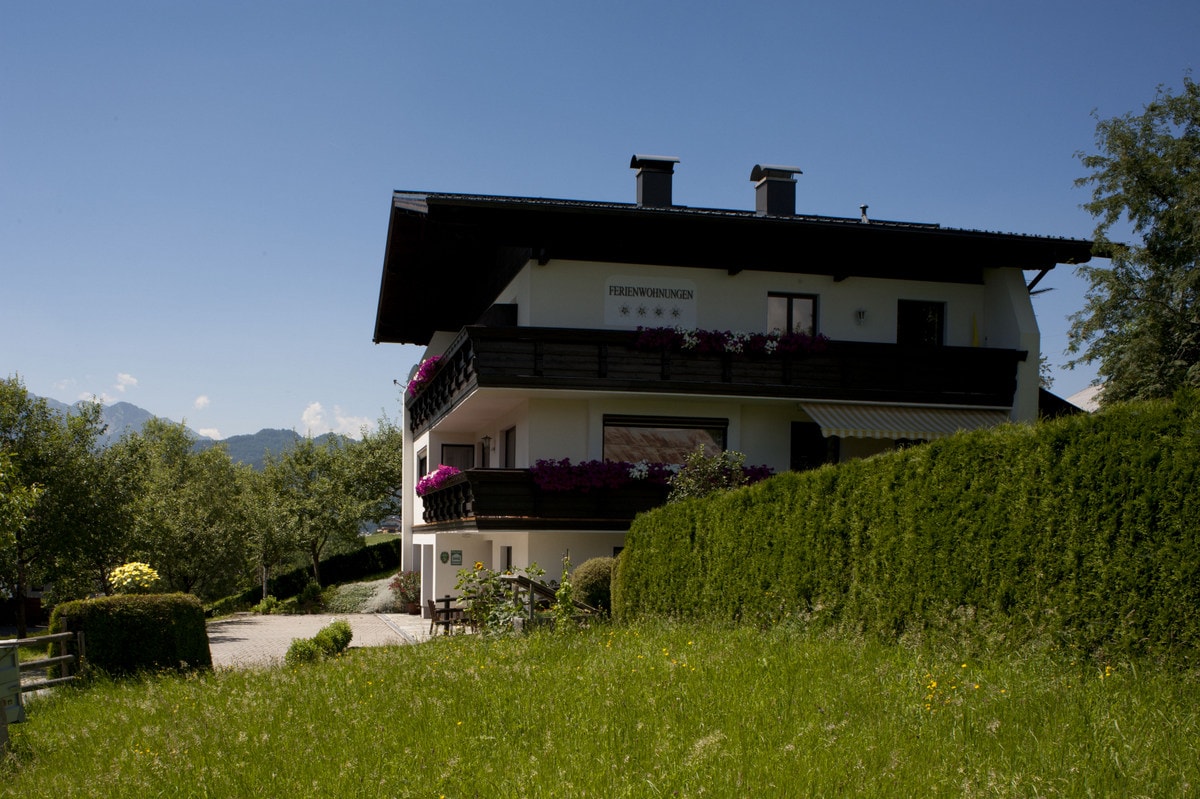 A multi-story building with a dark peaked roof is surrounded by lush greenery and colorful flower boxes on the balconies. The structure is complemented by a clear blue sky and distant mountains, offering a serene and natural setting.