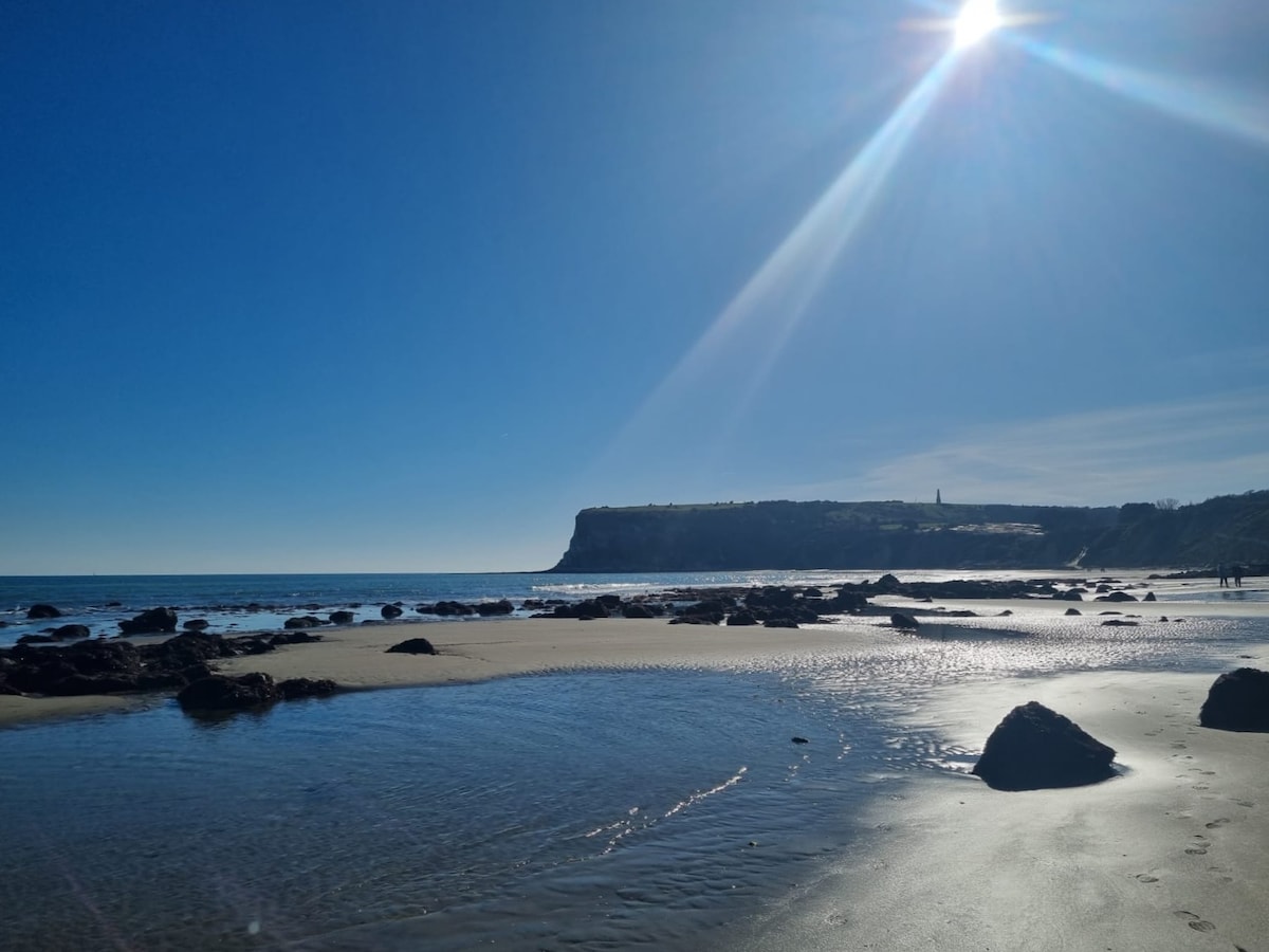 A sandy beach stretches along the coastline with clear blue skies overhead. Shimmering water reflects sunlight, creating a serene ambiance. Rocky formations are scattered across the shore, and a distant cliff visible in the background reveals natural beauty and inviting space for relaxation.