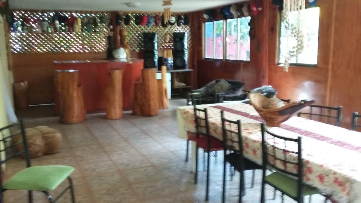 An inviting communal dining area features a long table with patterned tablecloth and an array of chairs. A wooden bar area with natural wood accents is visible in the background. Large windows allow natural light to fill the space, enhancing the warm atmosphere.