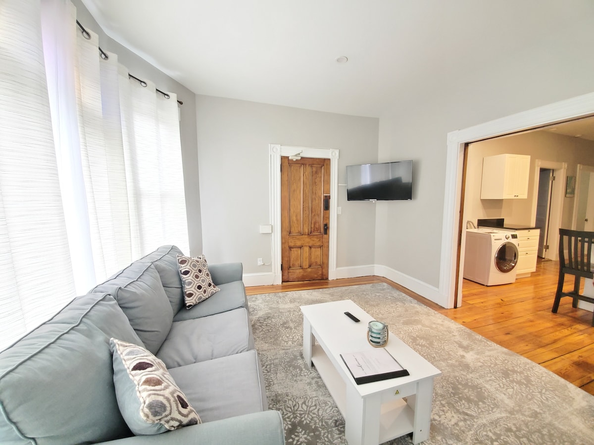 A cozy living room features a light grey sofa adorned with decorative pillows. A simple white coffee table sits in front, complemented by a flat-screen TV mounted on the wall. Natural light streams through large windows with sheer curtains, illuminating the space's warm wooden flooring.